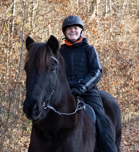 Edda sitter på en sort hest. Hun har lyst hår og rideklær på og smiler til kamera. Det er trær med høstfarger i bakgrunnen.