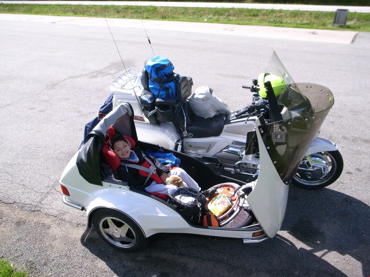 Leo sitter i sidevognen på en hvit motorsykkel. Han smiler til kamera og har flere leker med seg. Motorsykkelen står parkert på en asfaltvei.