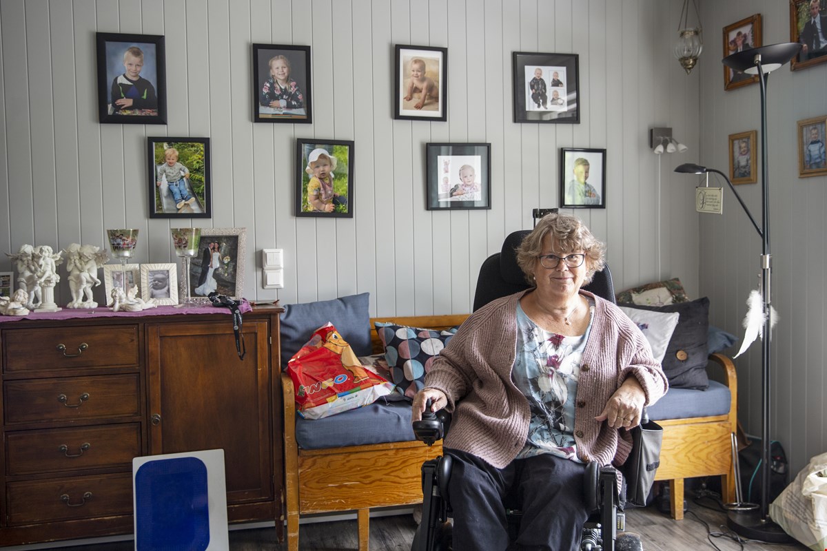 Portrett av Anne Karin foran en mørkerød trevegg. Hun har kort, grått  hår med fall i. Nakkestøtte til rullestol synes bak hodet, hun har på seg rosa strikkejakke med blomstrete topp under. Hun har på briller, perler i ørene og rundt halsen. Hun smiler til kamera.