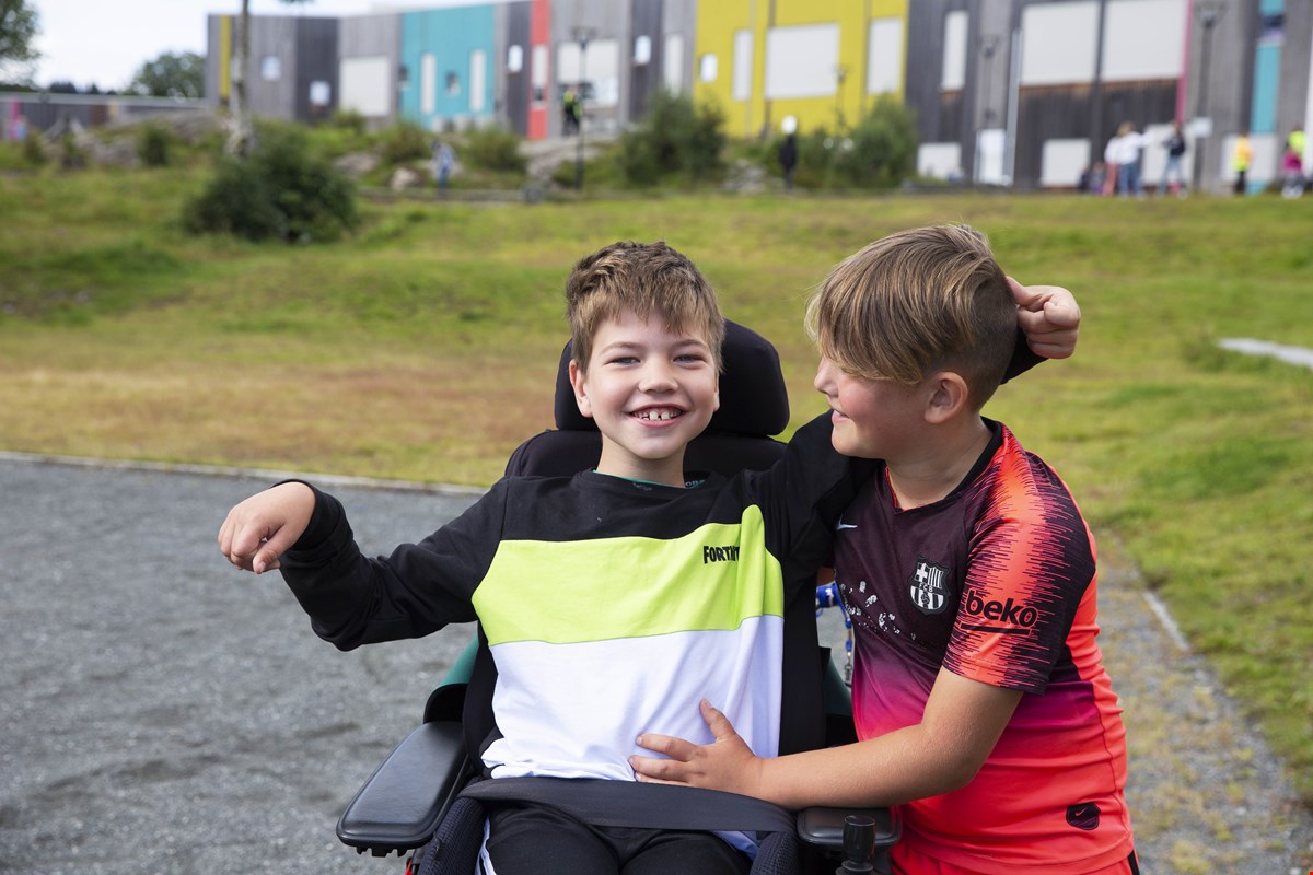 Even sitter i rullestolen og smiler til kamera. I bakgrunnen er et fargerikt skolebygg. Ved siden av han står en kamerat, som har armene delvis rundt og Even og smiler mens han ser på Even.