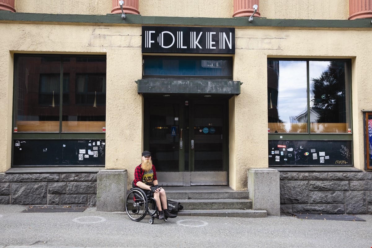 Helge sitter i manuell rullestolv ved trappen foran "Folken". Bygget er lysegult og over døren er det et sort skilt med hvite bokstaver som former "Folken". 