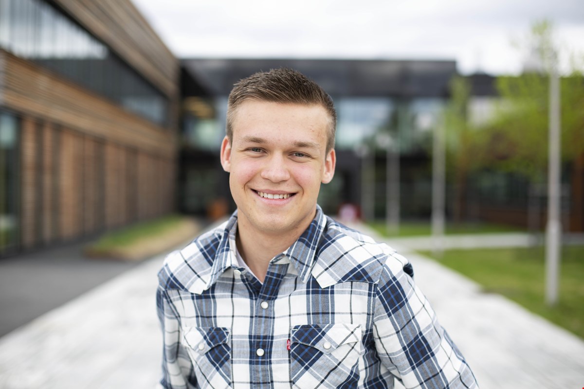 Nærbilde av Jon Eirik som smiler til kamera. Han har kortklipt, blondt hår og rutete skjorte i hvit og blå.  Skolebygget er uskarpt i bakgrunnen.