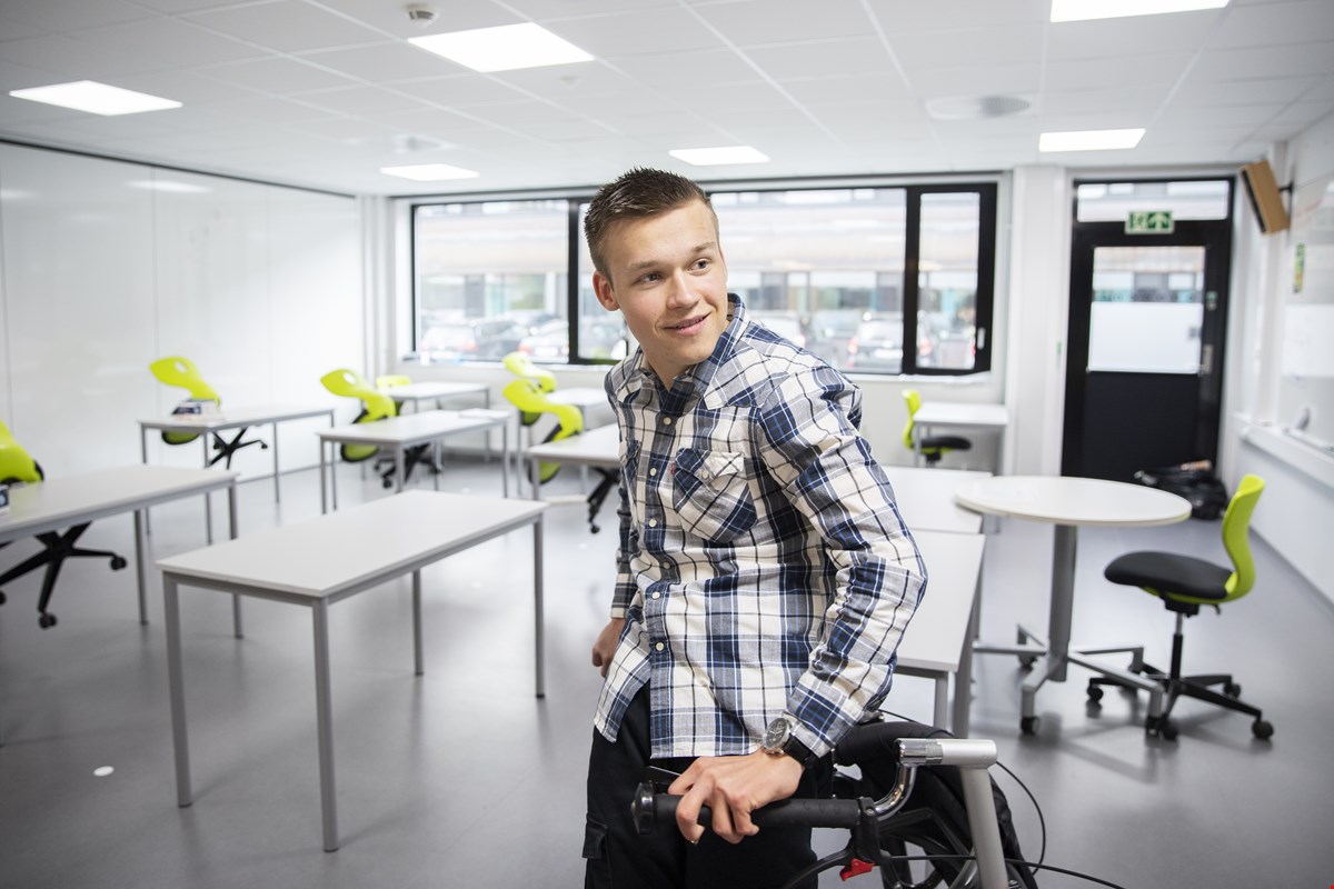 Jon Eirik står i klasserommet, ser til siden og smiler. Han har på seg rutete skjorte og går med rullator bak seg.