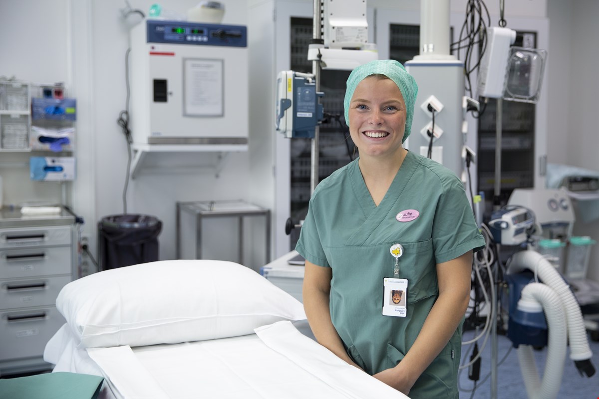 Julie står i en operasjonsstue på sykehuset og  har på seg grønn sykehusuniform. Hun har dekket håret med hårnett og smiler til kamera.