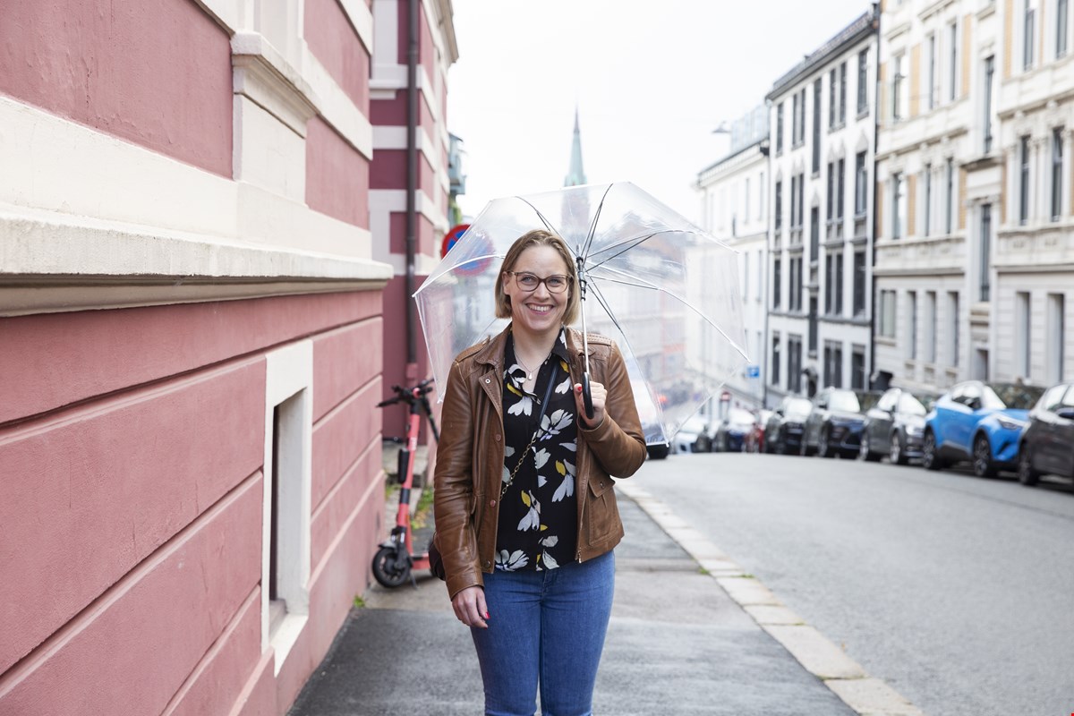 Kristine står i en gate i Oslo og smiler til kamera. Over skulderen holder hun en gjennomsiktig paraply.