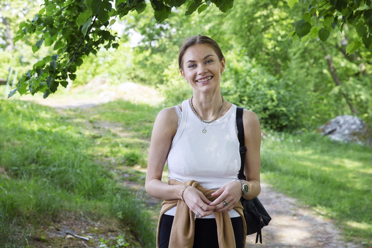 Marianne står i grønne omgivelser i en park. Hun har hvit singlet på og en brun genser rundt livet. Hun smiler til kamera.