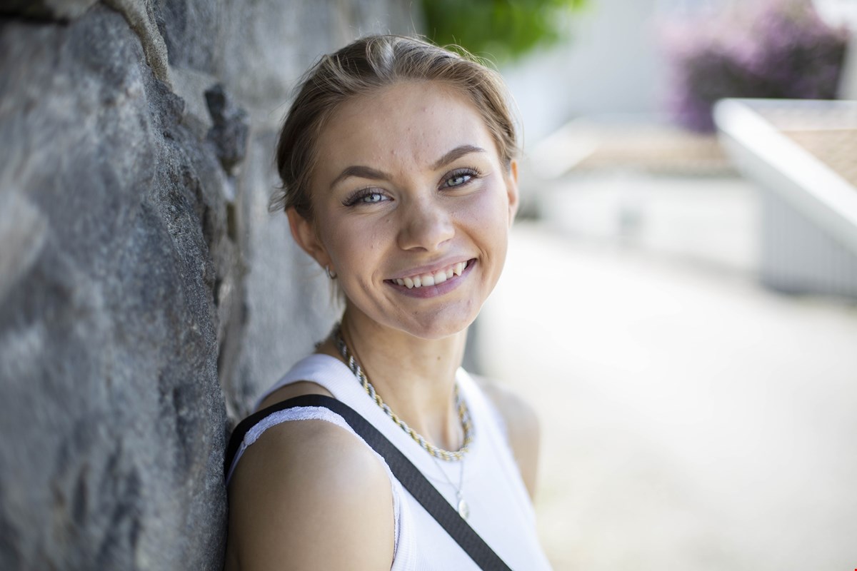 Nærbilde av Marianne som smiler mot kamera. Hun står inntil en mur, har på seg hvit singlet og oppsatt mørkeblondt hår.