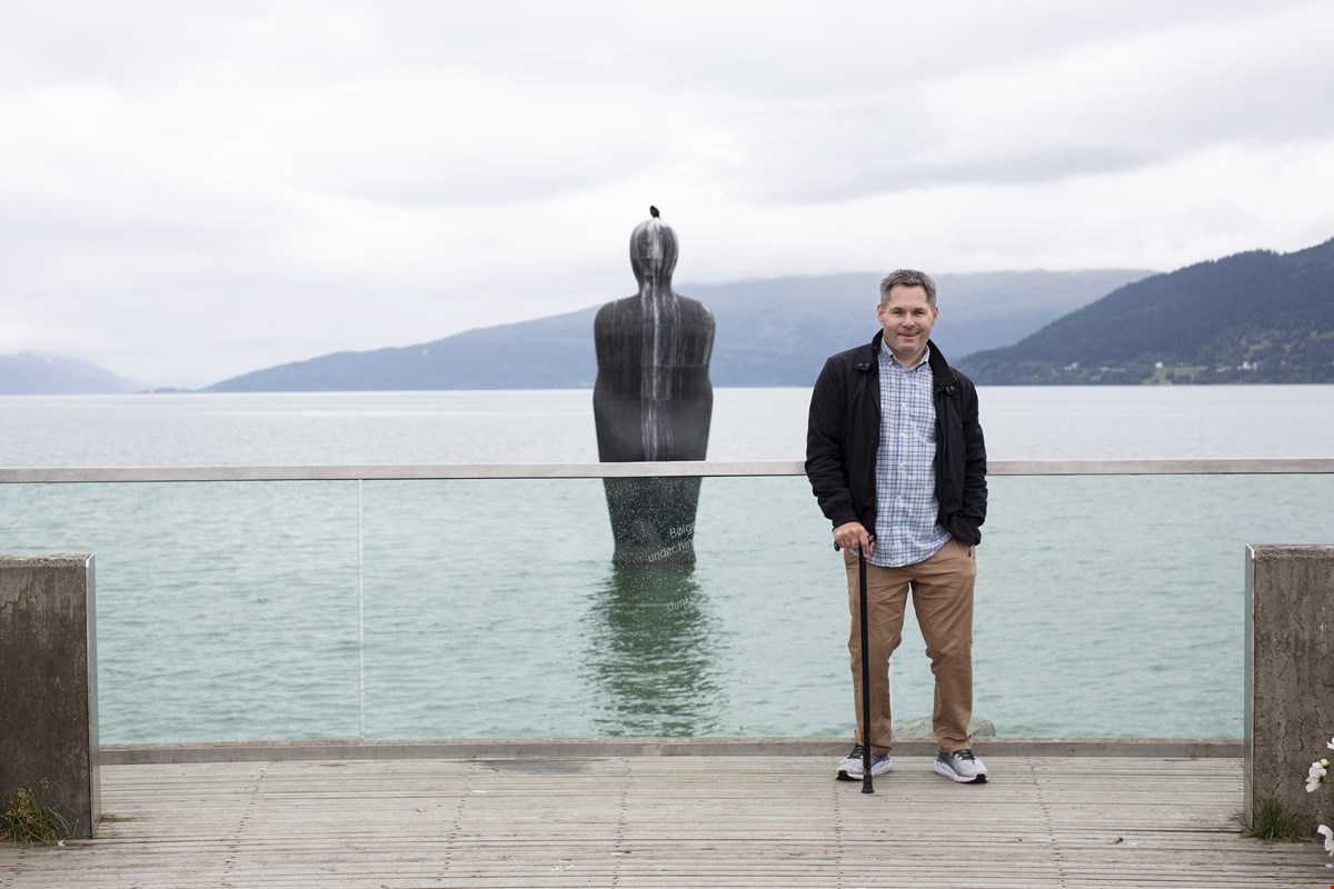 Tor Andreas står med sjøen i bakgrunnen. Midt i bildet er det en metallstatue som står i sjøen. Bildet viser Tor Andreas i helfigur, han støtter seg på en stokk og smiler til kamera.