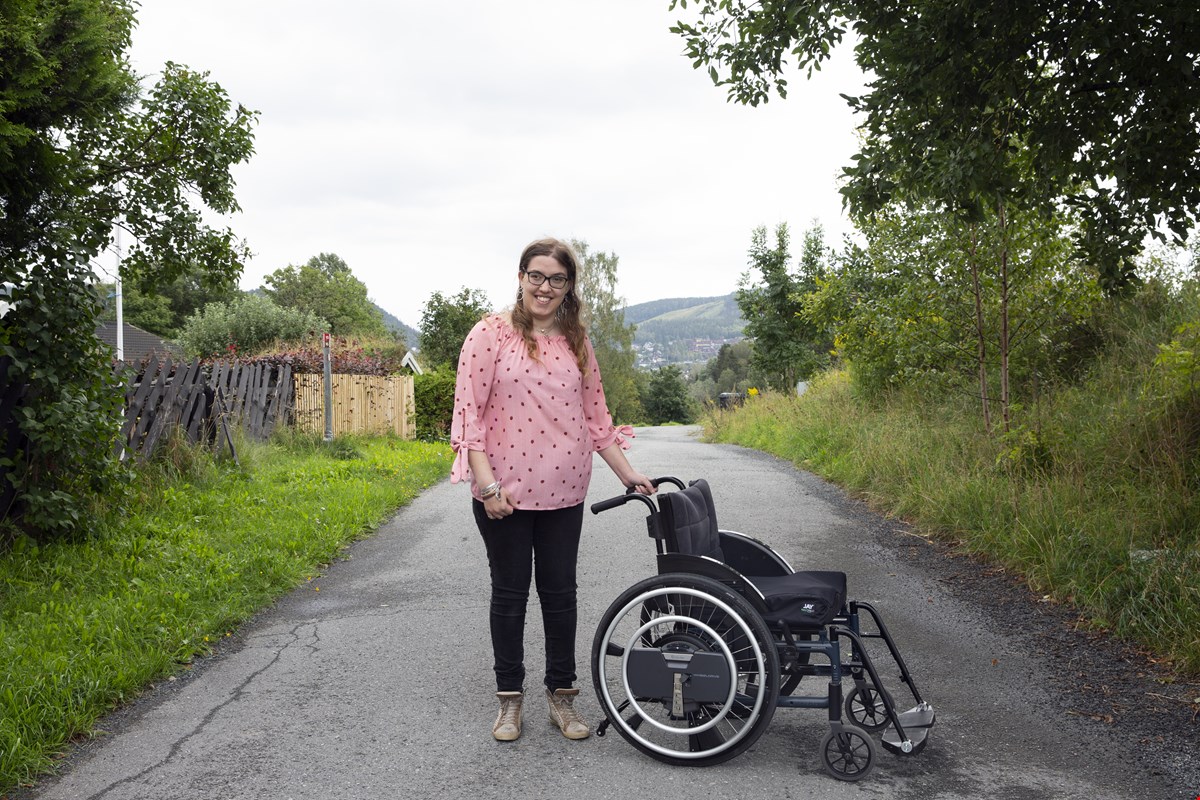 Victoria står ved siden av rullestolen sin og smiler til kamera. Rundt henne er det naturomgivelser. Hun har briller, mørkt, langt og krøllete hår og på seg har hun en ferskenfarget bluse med sorte prikker.