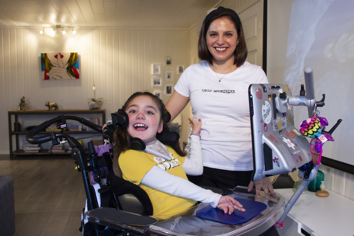 Isabella og Carolina står ved siden av hverandre og smiler til kamera. Isabella sitter i rullestol og har Tobii-skjermen foran seg. Bak dem er en lys stuevegg med bilder på.